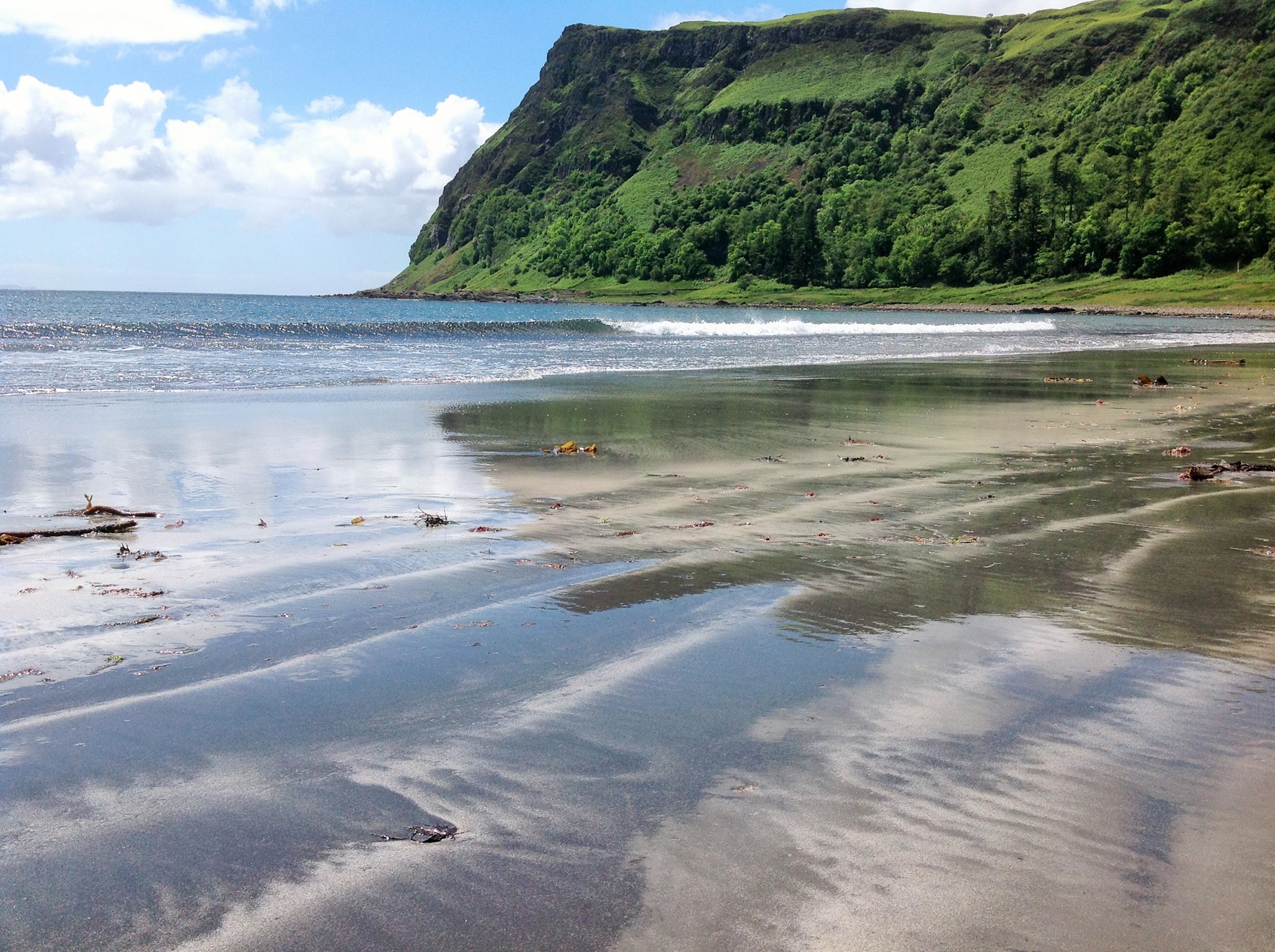 The grey sand beach at Carsaig, surrounded by a lush green headland and blue clouded skies as the waves lap on shore