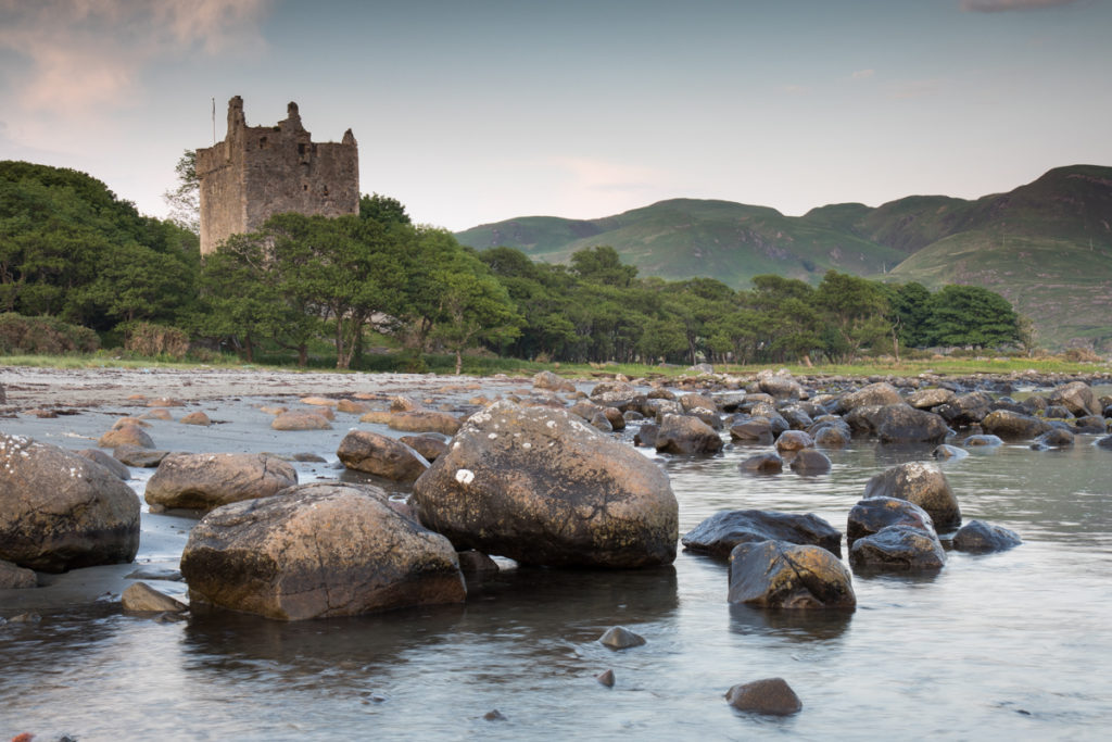 The ruins of Moy Castle poking out above the trees with a boulder strewn beach in front