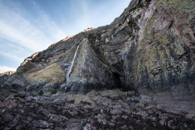 A walker heading across rocky coast to the entrance of the cave